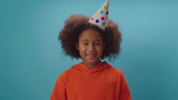 Menina afro-americana alegre em chapéu de festa de aniversário soprando cachimbo de laranja em pé sobre fundo azul. Criança feliz parabeniza com aniversário olhando para a câmera. — Vídeo de Stock