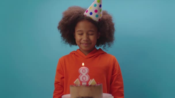 Afro-Amerikaans meisje met verjaardagshoed blaast in slow motion kaars nummer 8 uit. Acht jaar oud kind viert zijn verjaardag. Happy verjaardag meisje op blauwe achtergrond. — Stockvideo