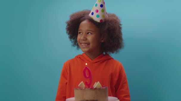 Menina afro-americana em chapéu de aniversário soprando vela número 9 em câmera lenta. A criança de nove anos celebra o aniversário. Feliz aniversário menina no fundo azul. — Vídeo de Stock
