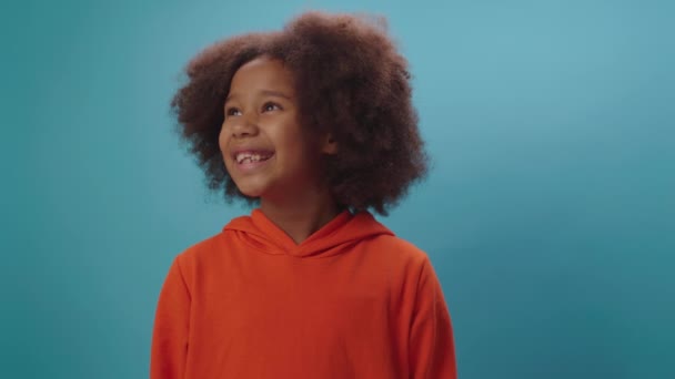 Lächelndes afroamerikanisches Schulmädchen in orangefarbener Kleidung, das in die Kamera blickt, die auf blauem Hintergrund steht. 7 Jahre altes Kinderporträt. — Stockvideo