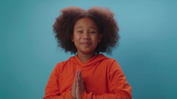 Une écolière afro-américaine plaidant debout sur fond bleu. Le gamin tenant la main pliée mendiant à propos de quelque chose regardant la caméra. Enfant demandant s'il vous plaît parler à la caméra. — Video