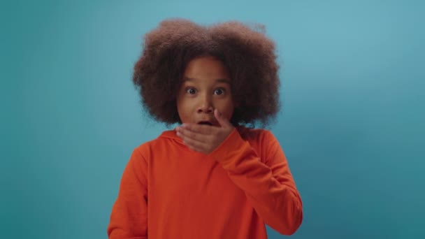 Miúdo chocado a abrir a boca e a fechá-la com a mão. Menina afro-americana mostrando choque ou wow emoção olhando para a câmera. Retrato de criança divertida no fundo azul. — Vídeo de Stock