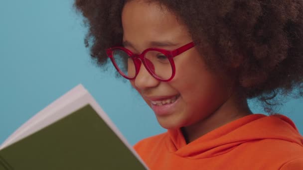 Gözlüklü Afrikalı Amerikalı kız kitap okuyor ve mavi arka planda duran kameraya gülümsüyor. Akıllı çocuk kitap okumayı sever.. — Stok video