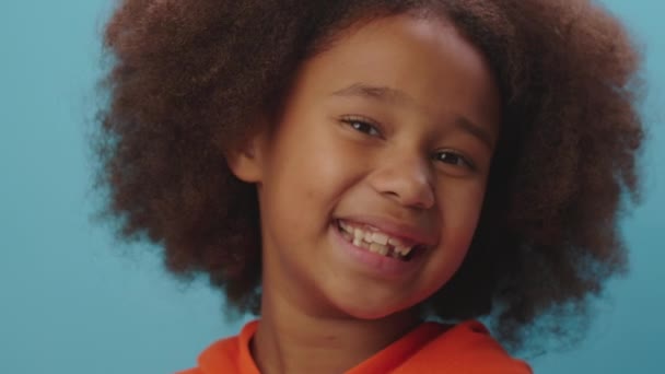 Portrait de fille afro-américaine souriante en vêtements orange regardant la caméra sur fond bleu. Gros plan sur un enfant heureux de 7 ans. — Video