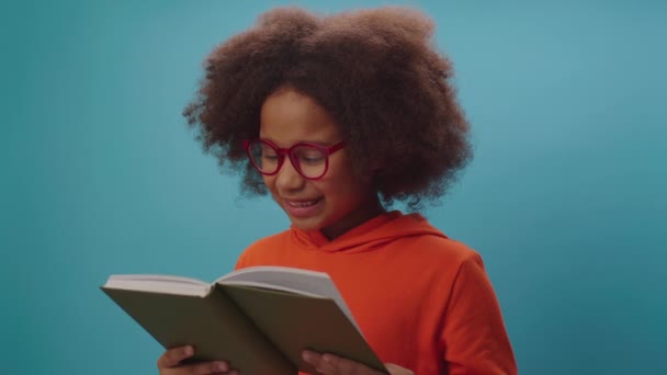 Afrikai amerikai iskolás lány szemüveg olvas könyvet és mosolyog a kamera előtt álló kék háttér. Okos gyerek szeret papírkönyveket olvasni..