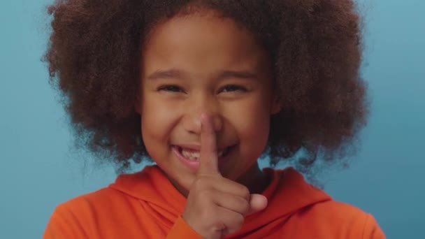 Закрывается симпатичная афроамериканская девушка, делающая жест молчания пальцем за палец на голубом фоне. Портрет ребенка, показывающий тишину и тихий знак пальцем. — стоковое видео
