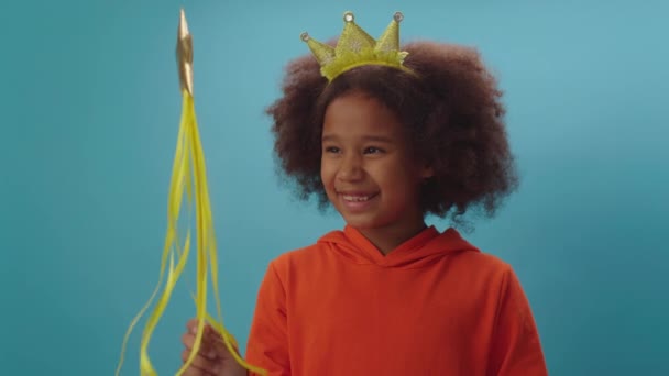 Całkiem pozytywne Afroamerykanka dziewczyna noszenie korony księżniczki i trzymając magiczną różdżkę uśmiechnięty patrząc na aparat stojący na niebieskim tle. — Wideo stockowe