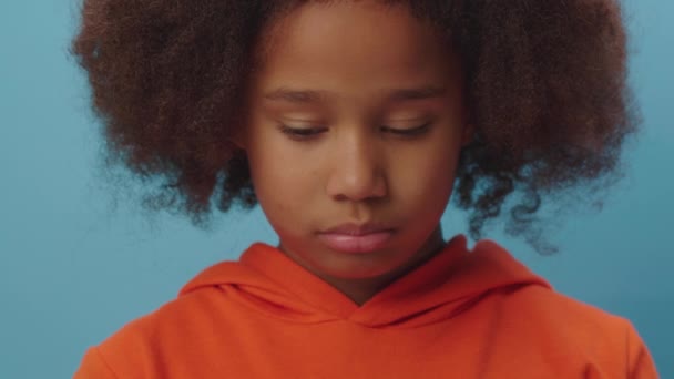 Close up de menina afro-americana chateada olhando para a câmera de pé no fundo azul. Criança mostra emoção infeliz. — Vídeo de Stock