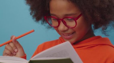 Gözlüklü Afrikalı Amerikalı kız kitap okuyor ve mavi arka planda duran kameraya gülümsüyor. Akıllı çocuk elinde kalemle kitap okumayı sever..