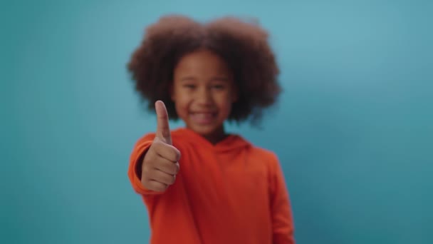 Nettes afroamerikanisches Mädchen zeigt lächelnd den Daumen in die Kamera, die auf blauem Hintergrund steht. Fokus verlagert sich vom Finger auf das Gesicht der Kinder. — Stockvideo