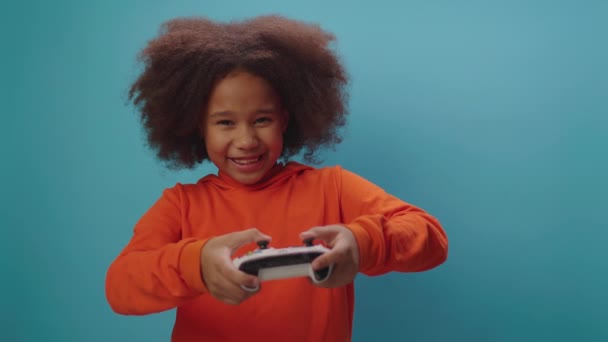 Aranyos afro-amerikai lány videojátékozik, kezében joystick kék háttérrel. A kölyök boldog, hogy megnyerheti videojáték. — Stock videók