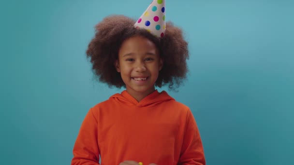 Wesoła Afroamerykanka w urodzinowym kapeluszu dmuchająca żółtą rurę stojąca na niebieskim tle. Happy kid gratuluje z urodzinami patrząc w kamerę. — Wideo stockowe