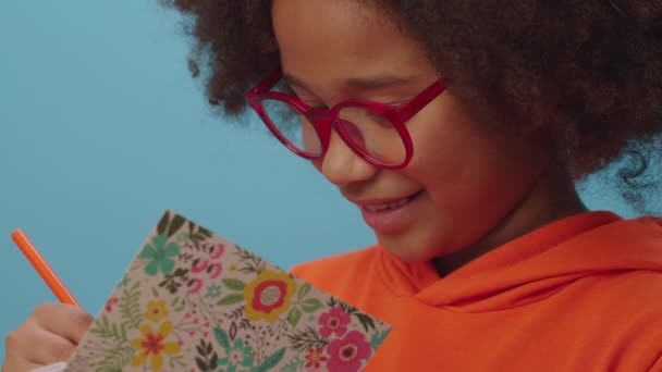 African American school girl in eye glasses writing with pencil in notebook and smiles at camera standing on blue background. Smart kid writing down the idea. — Stock Video