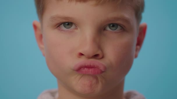 Nære på en søt gutt som viser uenighet stående på blå bakgrunn. Ungen er uenig ved å lage grimaser på leppene og riste hodet negativt.. – stockvideo