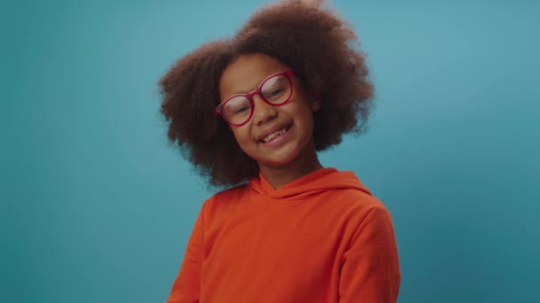 Fille afro-américaine souriante dans des lunettes de vue portant des vêtements orange regardant la caméra sur fond bleu. 7 ans enfant heureux dans des lunettes. — Video