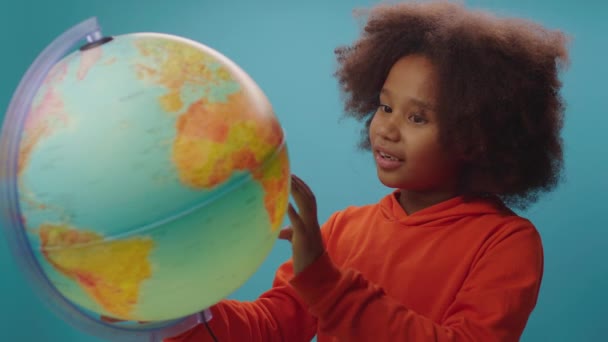 Цікава афроамериканська школярка, яка досліджує земну кулю, стоїть на синьому фоні. Освіта для дітей. — стокове відео