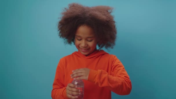 Красивая черная девушка пьет воду, держа пластиковую бутылку воды в руках, стоя на синем фоне. Ребенок утоляет жажду от питья водой. — стоковое видео