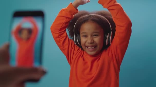 Ładna Afroamerykanka w słuchawkach tańcząca i śpiewająca patrząc na telefon komórkowy. Rodzice ręka za pomocą telefonu komórkowego nagrywania wideo z tańczącym dzieckiem. — Wideo stockowe