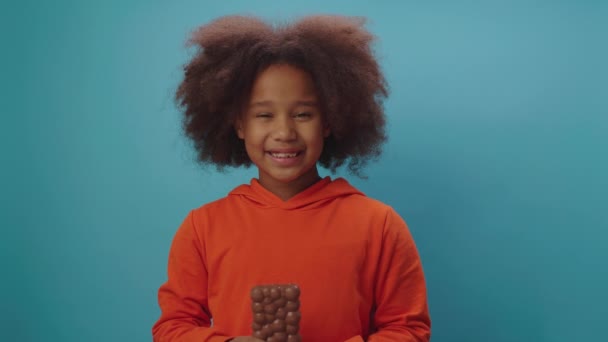 幸せな黒の少女手のカメラの青の背景に立って笑みを浮かべてチョコレートバーを保持食べている。かわいい子供はミルキーチョコレートの贅沢を楽しんで. — ストック動画