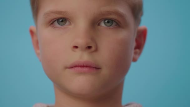 Портрет серьезного мальчика, смотрящего в камеру и улыбающегося на синем фоне. Закрыть 6-летнего ребенка с голубыми глазами. — стоковое видео