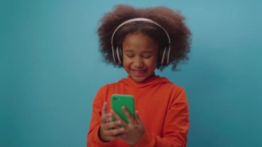 Kulaklık takan siyah bir kız mavi arka planda duran cep telefonuyla çevrimiçi video görüşmesi yapıyor..