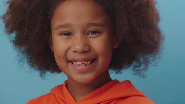 Portrait de fille afro-américaine en vêtements orange souriant à la caméra sur fond bleu. Gros plan sur un enfant heureux de 7 ans. — Video