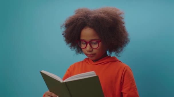 African American school girl in eye glasses reading book and making roof with book over her head looking at camera standing on blue background. Smart kid enjoys learning with paper books. — Stock Video