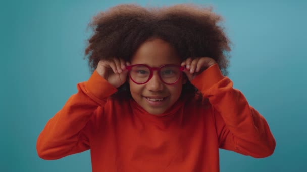Adorable African American girl in eye glasses smiling peering at camera. Happy kid in goggles intently looking at camera on blue background. — Stock Video