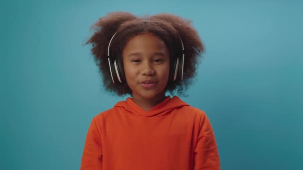Confident African American girl in wireless headphones talking at camera standing on blue background. Child blogger speaking to camera recording video. — Stockvideo