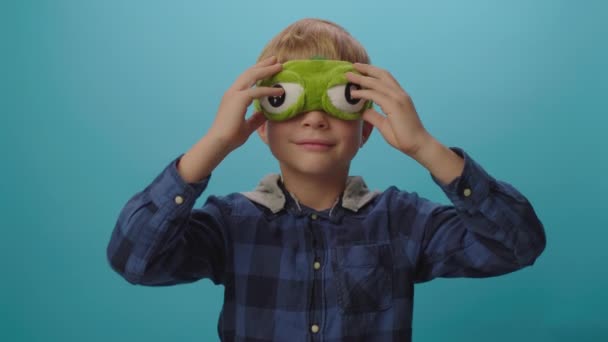 Niño rubio con divertida máscara de dormir con grandes ojos de pie sobre fondo azul. Hora de dormir con máscara. — Vídeos de Stock