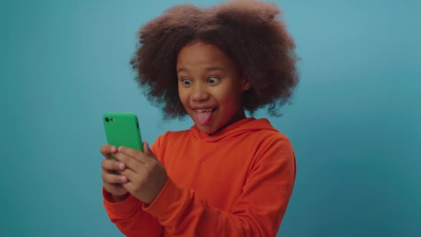 Afroamerykanka robi głupie miny patrząc na kamerę. Kid jest szczęśliwy, aby zabawne grymas i pokazać język za pomocą telefonu komórkowego. — Wideo stockowe