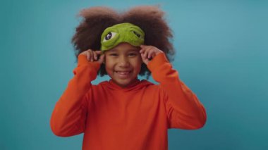 Mavi arka planda duran büyük gözlü, komik uyku maskesi takan sevimli Afro-Amerikalı kız. Maskeyle uyuma zamanı..