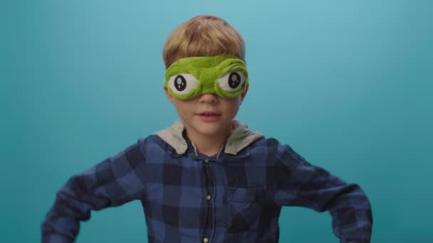 6 years old boy dancing and singing wearing funny frog mask standing on blue background. Kid wearing sleeping mask with big eyes. — Stock Video