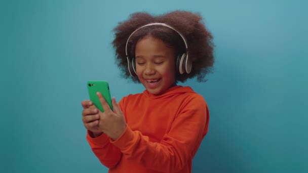 Hübsches schwarzes Mädchen mit Kopfhörern, das ein Lied singt und dabei auf das Handy auf blauem Hintergrund schaut. Lächelndes singendes Kind mit Handy. — Stockvideo