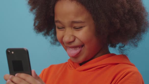 Close up of African American girl making silly faces looking at smartphone camera. Kid is happy to make funny grimaces and show tongue using cell phone. — Stock Footage Close up of African American girl making silly faces looking at smartphone camera. Kid is happy to make funny grimaces and show tongue using cell phone. Stock Footage