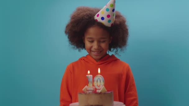 Menina afro-americana em chapéu de aniversário soprando vela número 10 em câmera lenta. A criança de dez anos celebra o aniversário. Feliz aniversário menina no fundo azul. — Vídeo de Stock