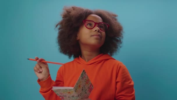 Pretty African American girl in eye glasses thinking holding notebook and pencil in her hands. Clever kid has an idea and writes down it in notebook. — Stockvideo