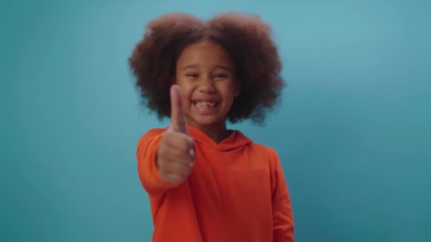 Мила афроамериканська дівчина, що показує великий палець, посміхається перед камерою, стоячи на синьому фоні. Фокус рухається від дитини до пальця. — стокове відео