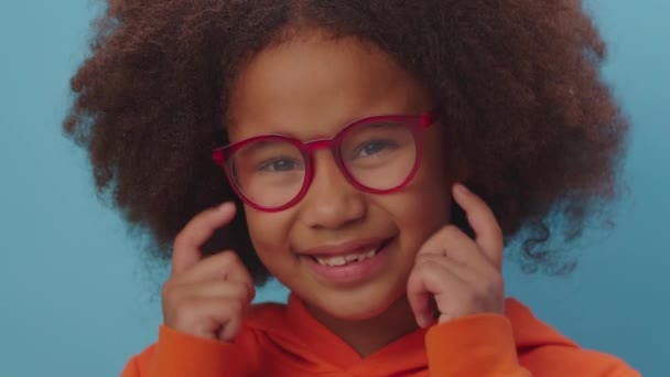 Portrét hezké černé dívky unavené nošením brýlí a třením očí stojících na modrém pozadí. Usmívající se dítě s problémy s viděním nosí brýle. — Stock video