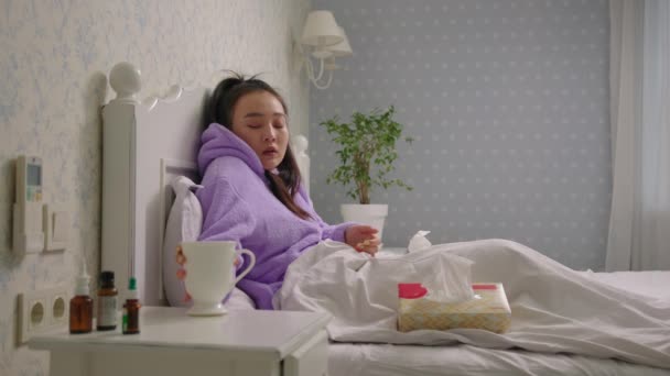 Νεαρή άρρωστη Ασιάτισσα που βήχει και φυσάει τη μύτη της με χαρτοπετσέτες στο κρεβάτι στο σπίτι. Γυναίκα πίνοντας ζεστό τσάι στο κρεβάτι. — Αρχείο Βίντεο