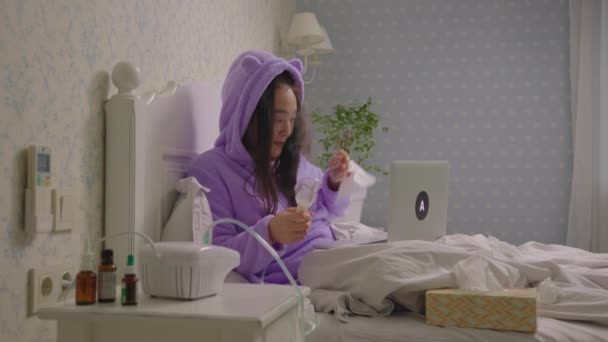 20s malade asiatique femme parler au médecin à l'aide d'un ordinateur portable tenant inhalateur dans les mains allongées dans le lit. Femme traitant la toux avec un inhalateur de médicament. Traitement de la grippe ou des virus respiratoires à domicile. — Video