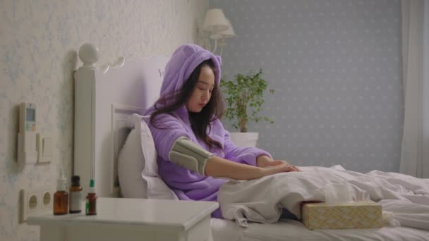 Femme asiatique malade mesurant sa tension artérielle à l'aide d'un tonomètre posé dans son lit à la maison. Femme a un mal de tête à cause de la pression artérielle. — Video