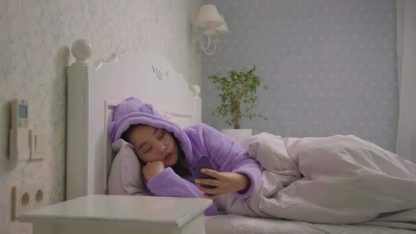 20-as évekbeli ázsiai nő lila pizsamában fekszik az ágyban mobiltelefonnal. Álmos nő böngészés online segítségével sejt az ágyban. — Stock videók