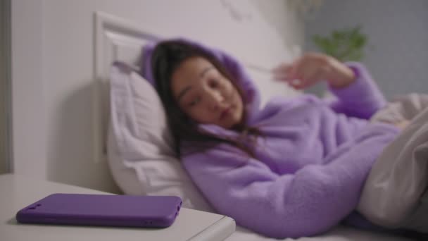 Primo piano di 20 anni donna asiatica in pigiama viola svegliarsi e prendere la cella in mano sdraiato a letto. Donna addormentata che utilizza il telefono cellulare al mattino. — Video Stock