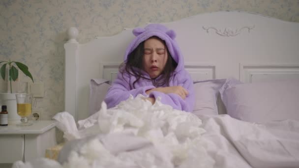 Húszas éveiben járó ázsiai nő köhög és kifújja az orrát szalvétával az ágyban. Influenza és egyéb légzőszervi betegségek otthon. — Stock videók