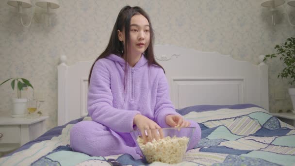 Mujer asiática bastante veinteañera viendo películas de miedo y comiendo palomitas de maíz sentada sola en la cama. Milenial mujer mira la televisión con palomitas de maíz en casa. — Vídeos de Stock