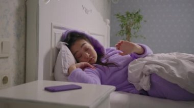 Mor pijamalı Asyalı bir kadın uyanıyor ve elinde cep telefonuyla yatakta yatıyor. Uykulu kadın sabahları cep telefonu kullanıyor..