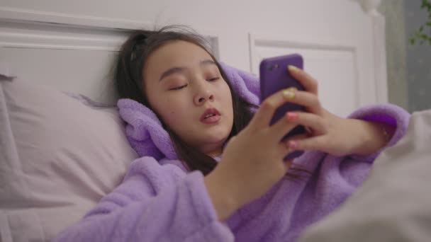 Тисячолітня азіатка друкує повідомлення за допомогою мобільного телефону, який лежить у ліжку. Щаслива молода жінка в піжамі користується мобільним телефоном. — стокове відео