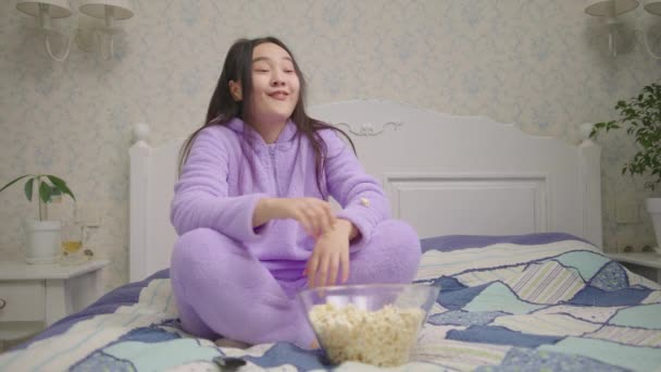 Asian woman watching funny movie or tv show and eating popcorn sitting alone on bed. Lady in purple pajamas laughing in front of tv. — Stockvideo