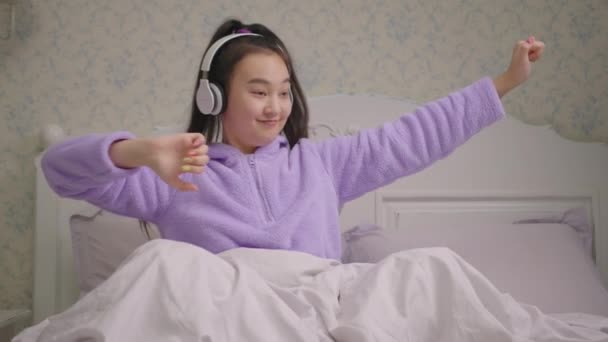 20-talets asiatiska kvinna njuter av att lyssna på musik i trådlösa hörlurar som sitter på sängen. Lycklig kvinna i lila pyjamas dansar med händerna. — Stockvideo